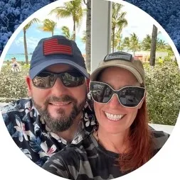 Smiling man and woman wearing sunglasses and caps with palm trees in the background outdoors.