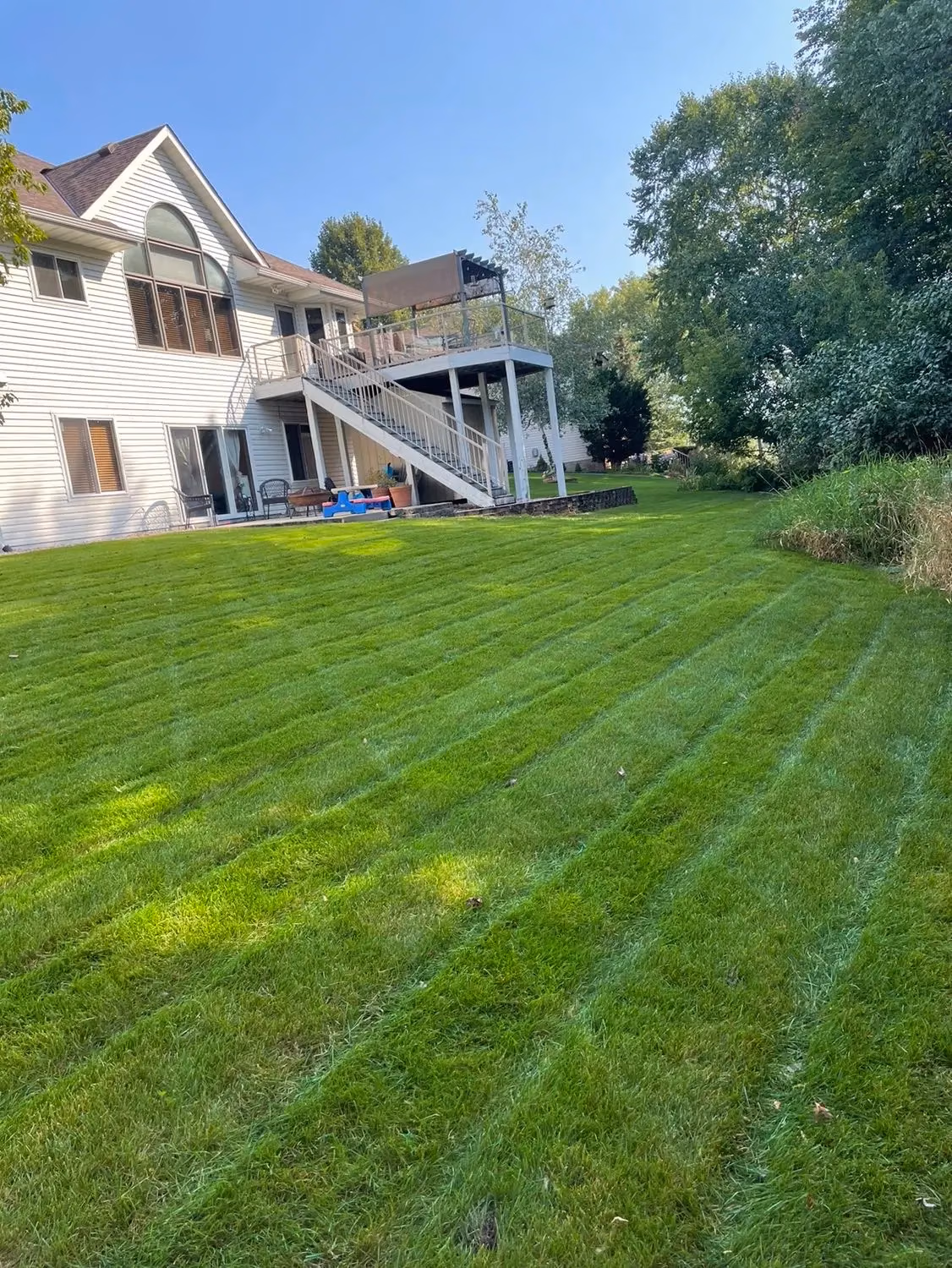 Large green lawn with fresh striped mowing in front of white two-story house with elevated deck and stairs.