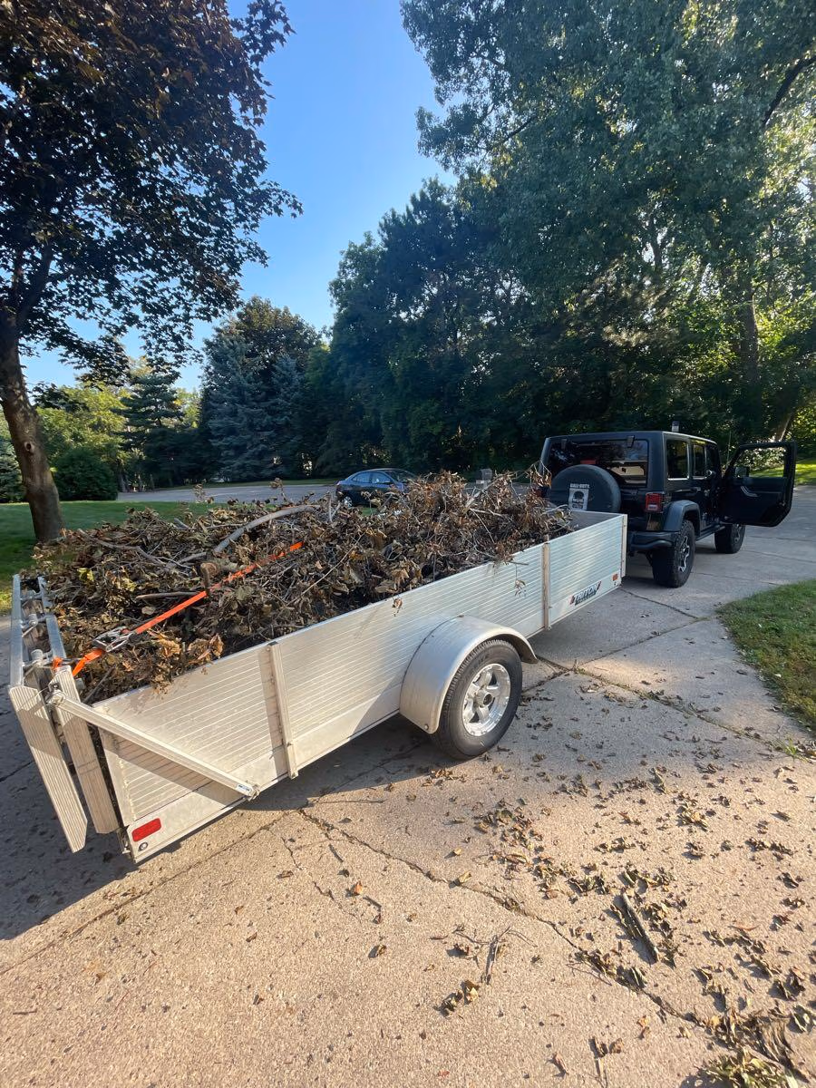 Utility trailer loaded with dry branches and leaves hooked to a black Jeep on a driveway.